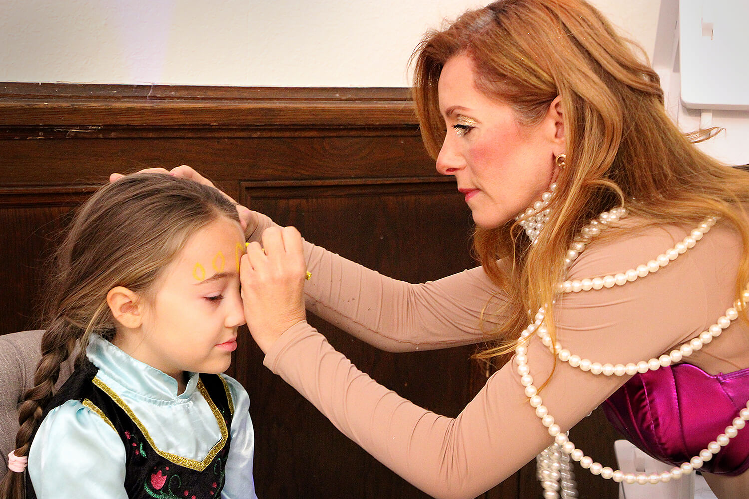  Karie Mathews wearing a purple top and layered pearl necklaces, is applying face paint to a child seated in a chair. The child is dressed in a costume with a light blue collar and black vest. The scene takes place indoors, with wooden paneling visible in