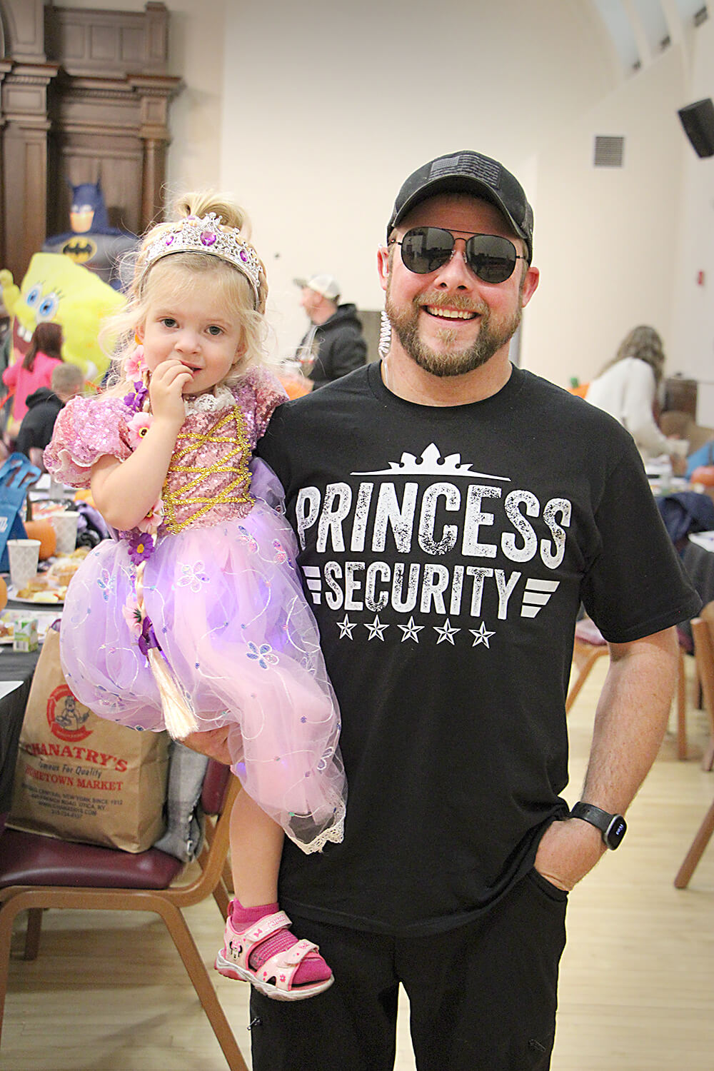 An adult wearing sunglasses and a black t-shirt labeled "PRINCESS SECURITY" holds a child dressed in a purple princess costume with a tiara. They are at an indoor event with other attendees and decorated tables visible in the background.