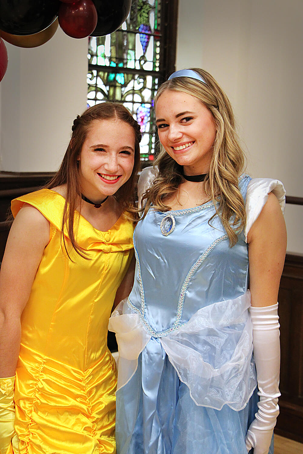 Two individuals dressed as princesses pose side by side indoors, smiling at the camera. One is wearing a yellow gown with a black choker, resembling Belle from Beauty and the Beast, while the other wears a light blue dress with white gloves and a headband