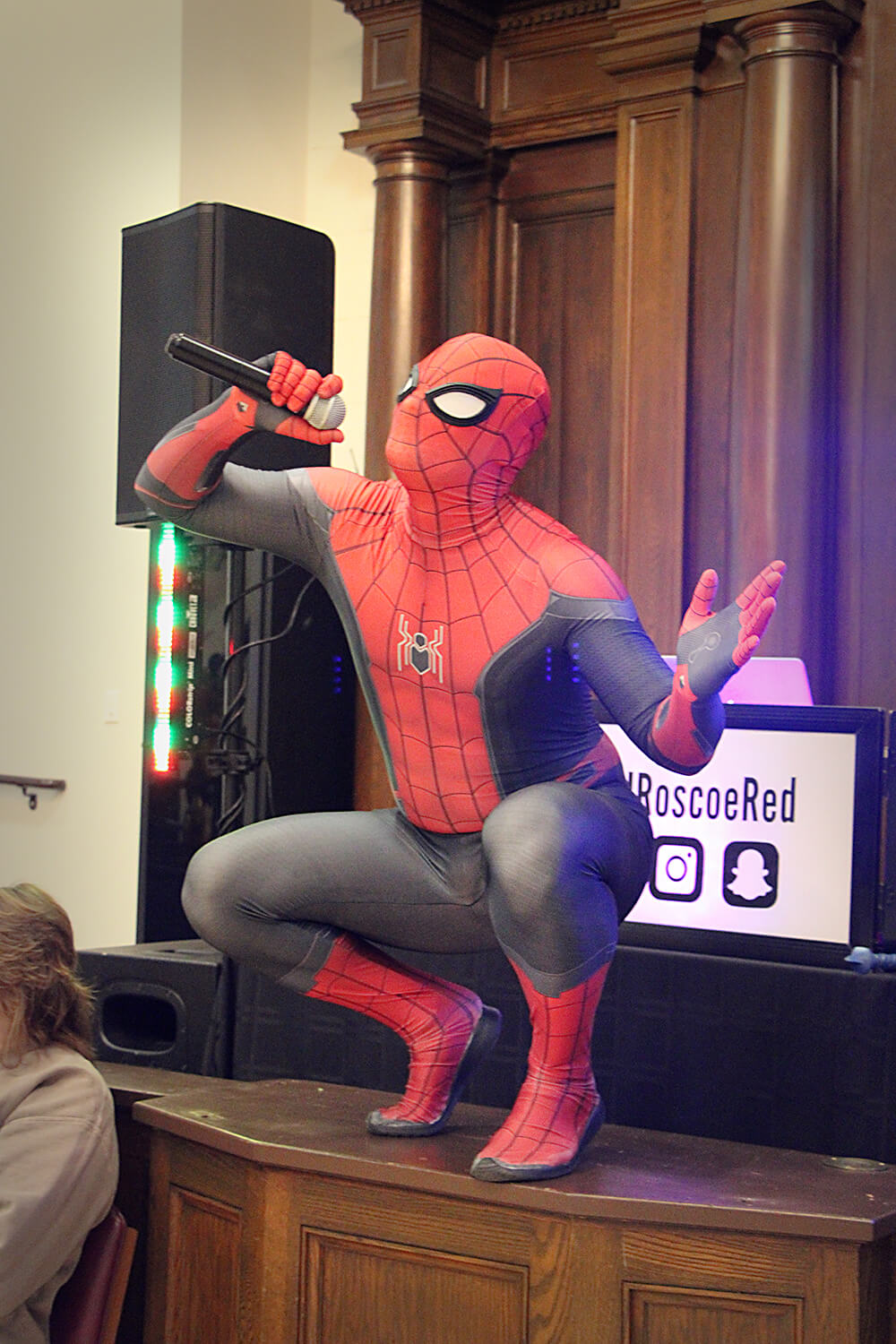 A person dressed in a full Spider-Man costume crouches on a wooden platform, holding a microphone to their mouth. The costume features red and blue colors with black web patterns and the Spider-Man logo. Behind them is a wooden structure with columns, and