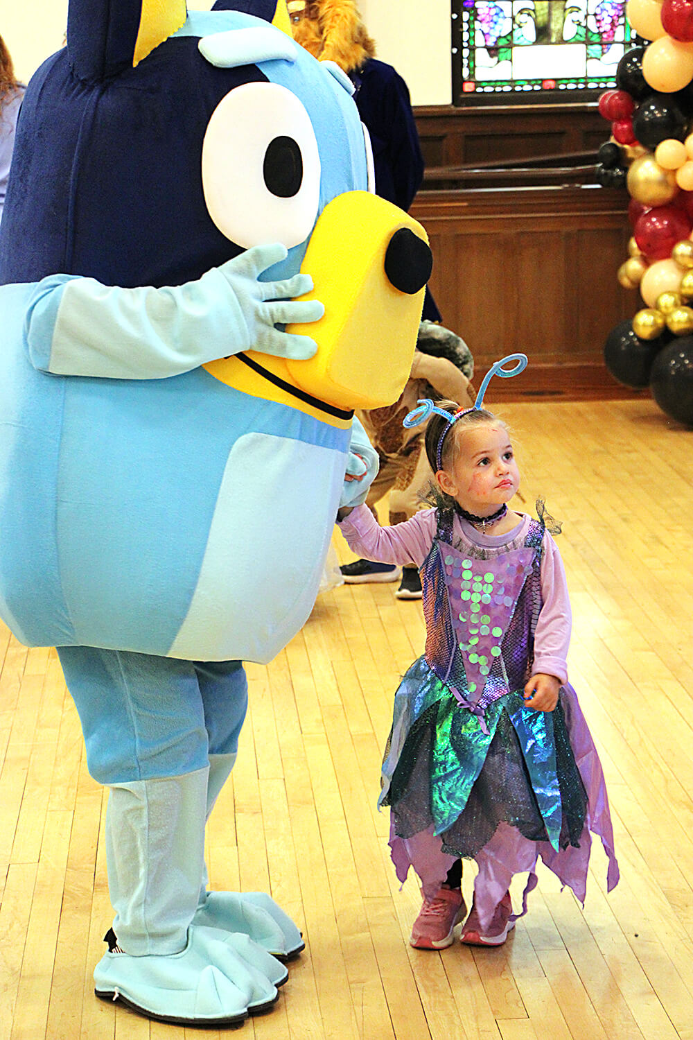 A person in a large blue cartoon character costume, Bluey, stands indoors on a wooden floor, holding hands with a young child dressed in a colorful insect or fairy-themed costume. The child looks up at the character. In the background, there are other att