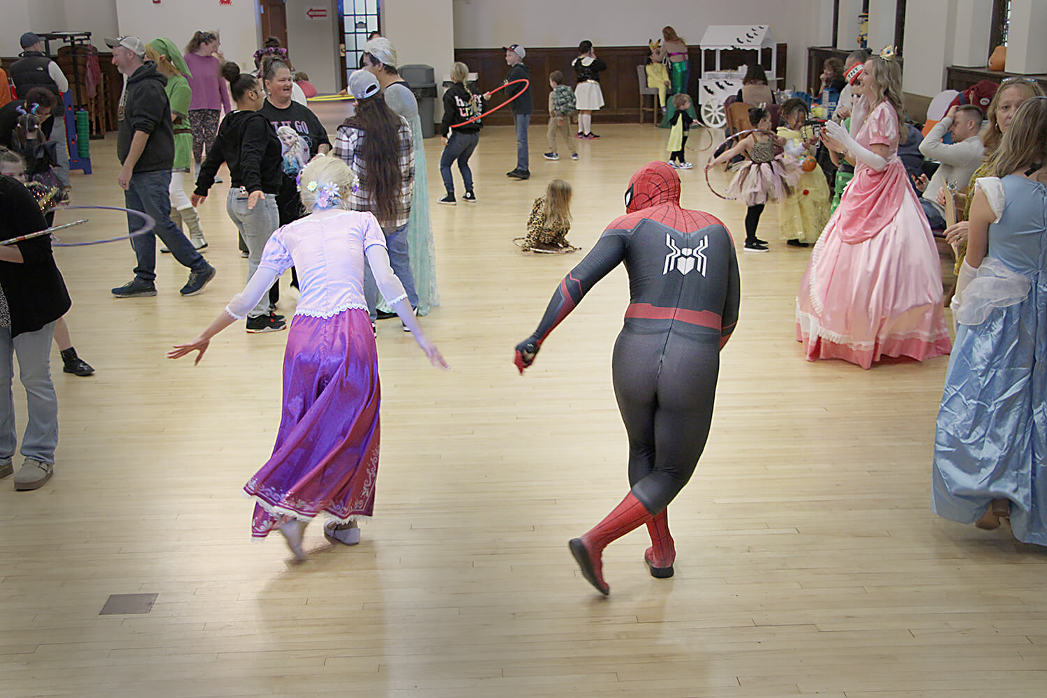 Two individuals dancing in the foreground — one dressed in a red and black Spider-Man costume, and the other in a long purple skirt, white top, and floral headpiece. The room has a wooden floor and is filled with other attendees in colorful costumes, incl