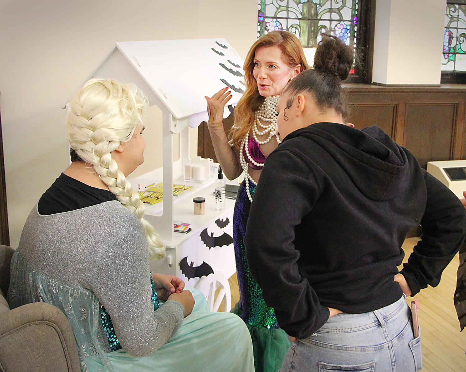 Three individuals are gathered at the RCIL Character Party. One is seated, dressed in a sparkly costume with a long braided wig. Another stands nearby in a mermaid-themed outfit with sequins and pearl accessories. The third person, dressed casually in a b