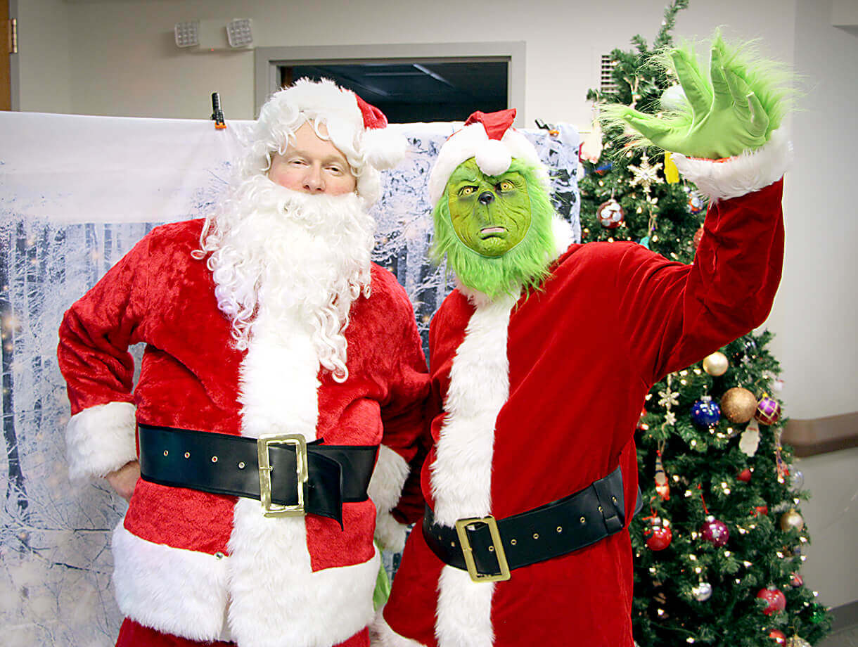 Two people dressed in festive costumes stand in front of a decorated Christmas tree. One is wearing a traditional Santa Claus outfit with a red suit, white trim, and a black belt. The other is dressed as the Grinch, also in a red Santa suit, with a green 